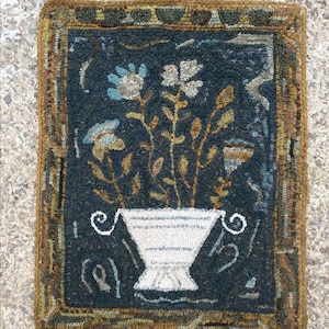 May include: A hand-woven rug with a dark blue background featuring a white vase with a floral arrangement. The vase has a striped pattern and the flowers are in shades of blue, white, and brown. The rug has a brown border with a geometric pattern.