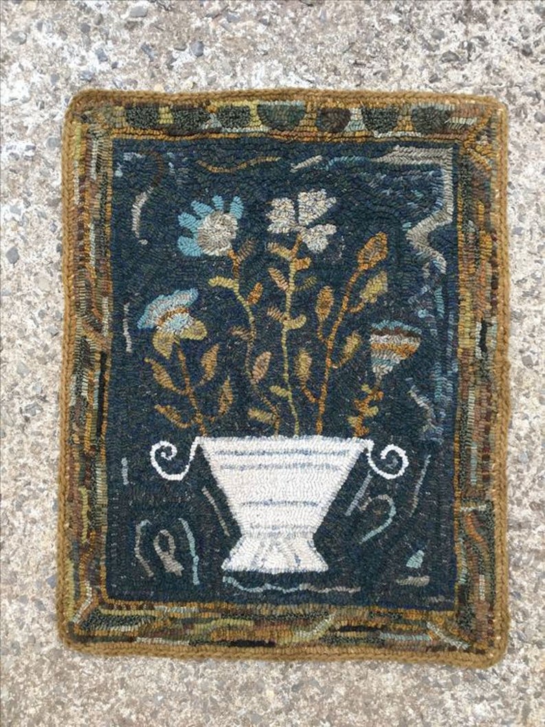 May include: A hand-woven rug with a dark blue background featuring a white vase with a floral arrangement. The vase has a striped pattern and the flowers are in shades of blue, white, and brown. The rug has a brown border with a geometric pattern.
