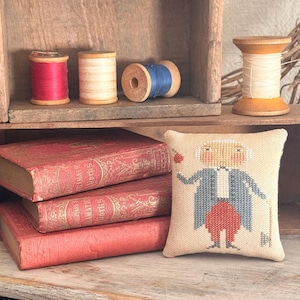 May include: A still life arrangement featuring vintage spools of thread in red, cream, and blue, alongside antique red books and a small cross-stitch pillow with a colonial figure. The items are arranged on a wooden shelf.