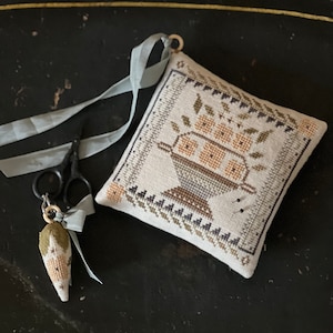 May include: A small, white, square pillow with a cross-stitch design of a bowl of fruit. The pillow is decorated with a blue ribbon and a small, green, cross-stitched ornament.