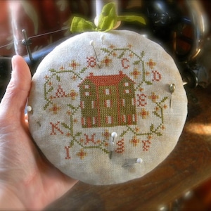 May include: A white fabric needlepoint with a green, brown, and orange design of a house with a red roof. The needlepoint is framed with a white border and has a few white pins in it.