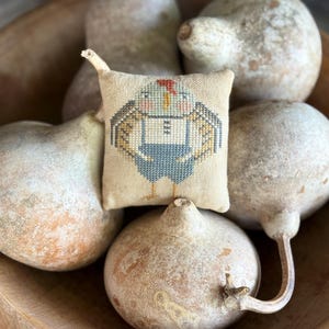 May include: A small, square, cream-colored fabric pincushion featuring a cross-stitched design of a stylized chicken wearing blue overalls. The chicken has a red comb, orange beak, and pink cheeks, with brown wings and legs.