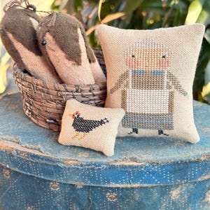 May include: A rustic still life features a woven basket holding three brown and tan fabric objects. Two small cross-stitch pillows depict a hen and a woman in an apron. The items rest on a blue patterned surface, evoking a farmhouse aesthetic.