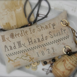 May include: A beige needlepoint pillow with the text "My Needle Is Sharp And My Hands Swift. 1871." The pillow is decorated with brown and white buttons and a wooden spool.