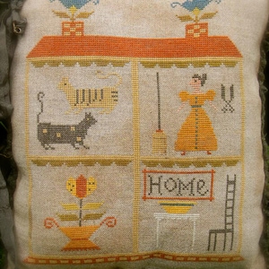 May include: A beige pillow with a cross-stitch design of a house with four rooms. The rooms depict a cat, a woman with a broom, a flower, and a ladder. The text "Home" is stitched above the ladder.