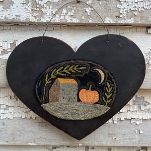 May include: A black heart-shaped wooden plaque with a needlepoint design of a house, a pumpkin, and a crescent moon. The design is in shades of green, brown, orange, and yellow.