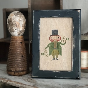 Op de afbeelding: Een ingelijste kruissteek van een grillige figuur met hoge hoed en groene jas, met takjes groen. De donkerblauwe lijst heeft een verweerde afwerking. Een decoratief eivormig object en een houten bijenkorf-achtig item staan op de achtergrond.