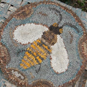 May include: A round, handmade rug featuring a stylized bee design. The bee has white wings, a yellow and black striped body, and brown and black details. The rug's background is gray and blue, with a brown border. The rug is placed on a blue woven surface.