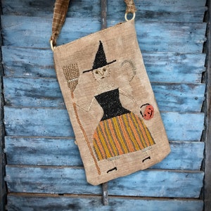 Pode incluir: Uma bolsa de tecido marrom com uma bruxa e uma vassoura bordadas. A bruxa está vestindo um vestido preto com listras laranja e amarelas. A bolsa tem uma alça marrom e uma fivela de plástico branca.