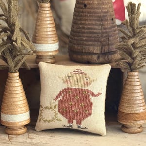 May include: A square beige fabric pillow featuring a cross-stitch design of a small figure in a red patterned outfit. The pillow is surrounded by three wooden cone-shaped decorative trees with brown fiber tops and light-colored thread accents.