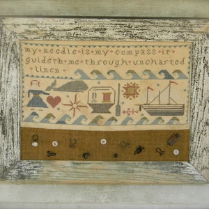 May include: Framed cross-stitch sampler with a nautical theme. The design includes a whale, sailboat, and waves, with the text "my needle is my compass it guideth me through uncharted linen." The frame is distressed white.
