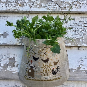 May include: A beige linen wall hanging with a cross-stitch design of a woman and chickens. The wall hanging is filled with green plants and hanging from a wire hook on a white weathered wood wall.