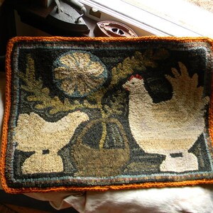 May include: A hand-stitched rug with an orange border features two white chickens with brown eyes and a floral design in the background.
