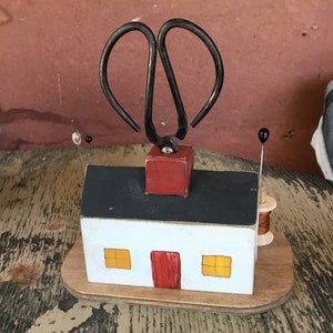 May include: A white wooden house with a black roof and red door. The house has two yellow windows and a red chimney. A pair of black metal scissors is attached to the top of the chimney. The house is sitting on a wooden base.