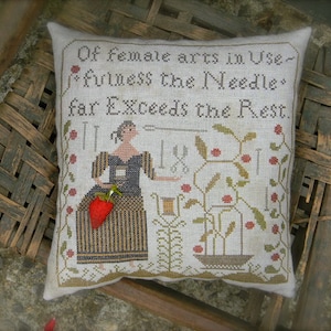 May include: A white pillow with a cross-stitch design featuring a woman holding a strawberry and the text "Of female arts in Use-fulness the Needle far Exceeds the Rest."