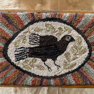 Puede incluir: Un pájaro negro con un ojo blanco se representa en un tapiz de crochet. El pájaro está en el centro de un círculo con un fondo blanco y un borde rayado marrón, verde y naranja.