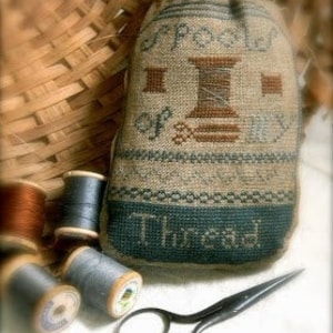 May include: A small, brown and blue fabric pouch with the words "Spools of Thread" embroidered on it. The pouch is filled with spools of thread and is sitting on a white surface next to a pair of scissors.