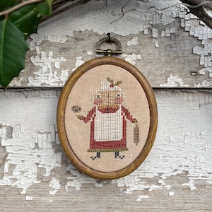 May include: A framed cross-stitch of a woman in a red dress and white apron holding a small bird. The woman has a red and green holly sprig in her hair. The frame is brown wood with a brass hook.