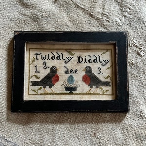 Puede incluir: Un bordado en punto de cruz enmarcado con dos pájaros negros con pecho rojo, un nido con un huevo azul y el texto "Twiddly Diddly Dee". Los números 1, 2 y 3 están bordados. El marco negro tiene un acabado envejecido.
