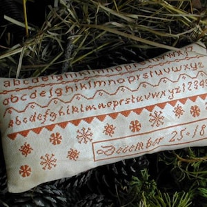 May include: A white pillow with an orange embroidered alphabet and snowflake design. The pillow reads "December 25, 1865".