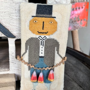 Puede incluir: Una almohada de punto de cruz con un personaje caprichoso con cara de calabaza que lleva un sombrero de copa y sostiene una cuerda de maíz dulce. El personaje está vestido con una chaqueta gris, pantalones azules y una camisa blanca con una corbata de lazo negra.