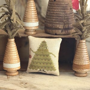 May include: A square beige fabric pincushion featuring a cross-stitched green tree with a face and a small red ornament, surrounded by four wooden spool-shaped decorative trees with textured green fiber tops and wrapped thread.