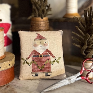 May include: A small, square, beige pillow with a cross-stitched Santa Claus design. The Santa figure is wearing a red suit and hat, with a black belt and boots. He is holding green sprigs. A pair of scissors with red handles rests nearby.