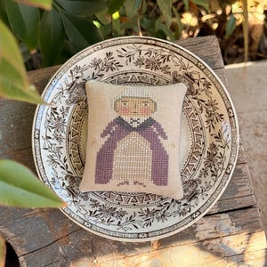 Può includere: Un piccolo cuscino quadrato beige a punto croce con una figura con un copricapo bianco e vesti viola. Il cuscino poggia su un piatto decorativo con un motivo floreale. Il piatto è su una superficie di legno stagionato.