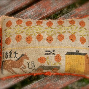 May include: A beige pillow with an embroidered design of a horse pulling a wagon with a house on it. The wagon is labeled "1824 LB". The house is yellow and has a red roof. The pillow has a red border and a fringe.