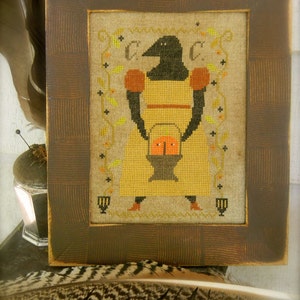 May include: A framed cross-stitch design featuring a black crow holding a basket of orange pumpkins. The crow is wearing a yellow dress with red sleeves. The background is a beige linen fabric with a green vine pattern.