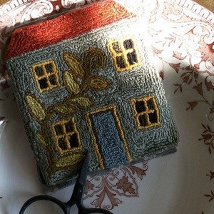 May include: A hand-stitched needlepoint house with a green roof, orange trim, and a yellow vine with leaves. The house has three windows and a blue door.