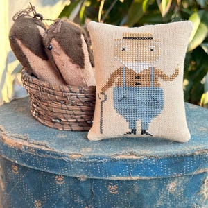 May include: A small, beige cross-stitch pillow featuring a man with a hat, mustache, and overalls, holding a cane. A woven basket with two brown, fabric objects sits behind the pillow. The objects are on a blue, patterned surface.