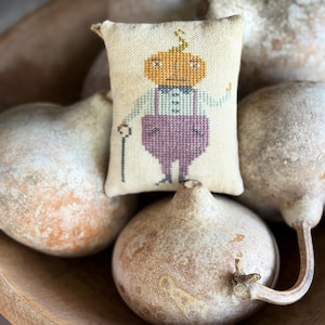 May include: A small, rectangular, cream-colored fabric pillow featuring a cross-stitched illustration of a pumpkin-headed figure wearing a light blue shirt and purple trousers, holding a thin black cane in one hand and standing upright.