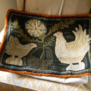 May include: A hand-stitched rug with a dark background featuring two white chickens and a green leafy plant. The rug has an orange border and is displayed on a white and orange patterned chair.