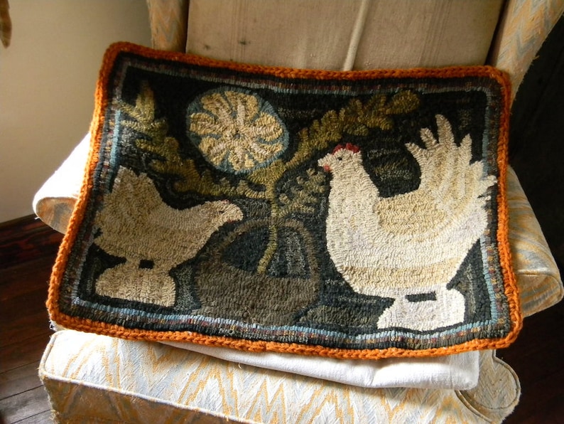May include: A hand-stitched rug with a dark background featuring two white chickens and a green leafy plant. The rug has an orange border and is displayed on a white and orange patterned chair.