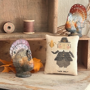 May include: Thanksgiving-themed display featuring a cross-stitched pillow with a pilgrim figure, two turkey figurines, and spools of thread. The color palette includes beige, brown, orange, and gray. The items are arranged on a rustic wooden shelf, evoking a vintage aesthetic.