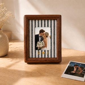 Puede incluir: Un marco de fotos rectangular de madera marrón oscuro con un paspartú de rayas verticales blancas y negras. El paspartú muestra las letras doradas R y J separadas por un símbolo de corazón. Una foto de una pareja está centrada.