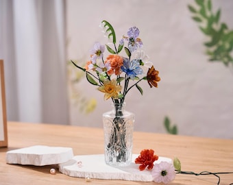 May include: A clear glass vase with a textured pattern holds a bouquet of multi-colored glass flowers, including orange, blue, purple, and white blossoms with green stems. Two loose glass flowers and small white pearls lie on the surface nearby.