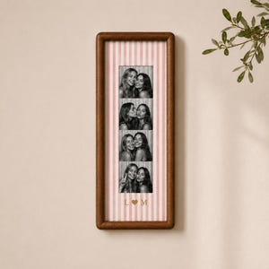 May include: A vertical rectangular wooden frame containing a strip of four black and white photographs of two people. The background inside the frame features light pink and white vertical stripes, with the letters L and M separated by a heart.