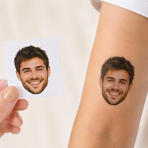 May include: A temporary tattoo featuring a realistic color portrait of a smiling person with dark hair and a beard, shown applied to skin alongside a square white paper card displaying the identical facial image for comparison and reference.