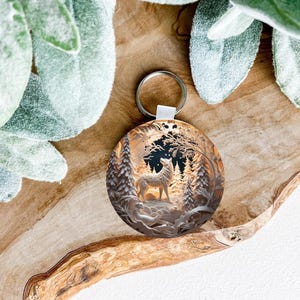 May include: A round, silver keychain with a detailed image of a wolf howling in a snowy forest. The keychain is on a wooden surface.