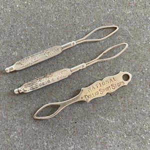 May include: Three vintage silver collar button hook tools. The first is labelled "The New Style Collars". The second is labelled "Derge-Dodenhausen Clothiers & Co.". The third is labelled "National Dollar Shirt Shops".