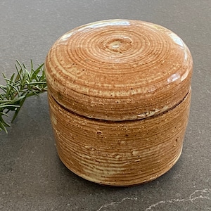 May include: A small, round, ceramic jar with a lid. The jar is a light brown color with a white glaze. The lid has a slightly raised center and is decorated with a swirling pattern.