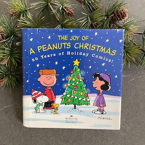May include: A square book titled "THE JOY OF A PEANUTS CHRISTMAS" with illustrations of Charlie Brown, Snoopy, and Lucy around a Christmas tree. The cover is blue with white stars and the Hallmark Books logo.