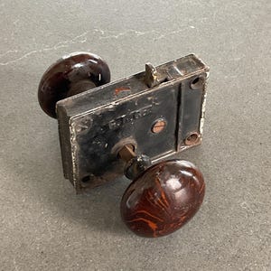 Puede incluir: Cerradura de puerta antigua negra con dos pomos de cerámica marrón. La cerradura tiene forma cuadrada y está hecha de metal. Los pomos tienen un patrón de espiral.