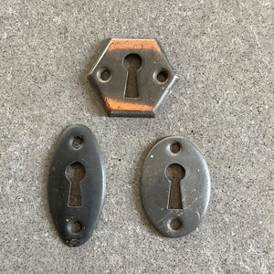 May include: Three metal keyhole escutcheons. One is a hexagonal plate with a weathered orange and gray finish, featuring two screw holes. The other two are oval-shaped plates in a matte gray finish, each with two screw holes for mounting.