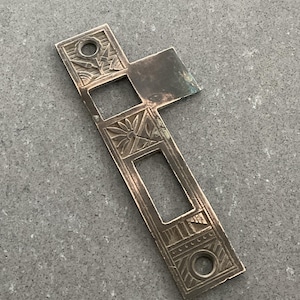 May include: A silver-colored, rectangular metal object with an ornate design. The object has a cross-like shape with cut-out sections and decorative floral and geometric patterns. The surface shows signs of wear, suggesting it is an antique or vintage piece.