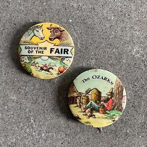 May include: Two vintage souvenir buttons. The first button is yellow with a white horse, a brown cow, and the text "Souvenir of the Fair". The second button is yellow with a man sitting on a log, a dog, and the text "The Ozarks".
