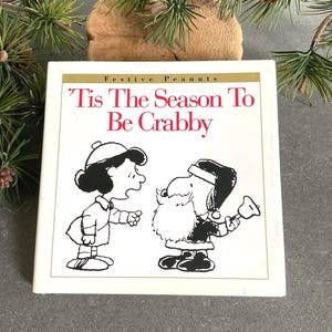 May include: A white square book with the text "'Tis The Season To Be Crabby" in red. The cover features black and white illustrations of Peanuts characters Lucy and Snoopy in a Santa Claus outfit. The book is part of the "Festive Peanuts" collection.
