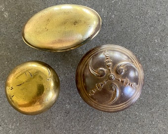 May include: Three vintage brass trinket boxes. One box is oval shaped with a smooth surface. The second box is round with a floral design. The third box is round with a smooth surface.